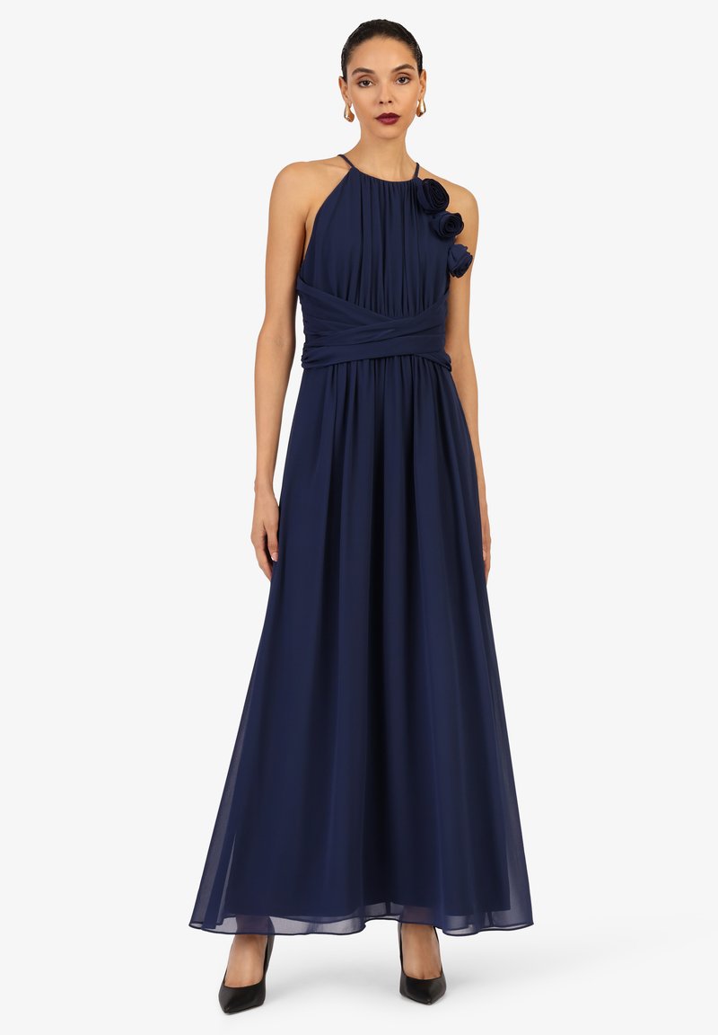 Langes, navyblaues Kleid aus Chiffon mit plissiertem Oberteil, Halterneck und dekorativen Stoffblumen auf einer Schulter. Sanfte, fließende Silhouette.
