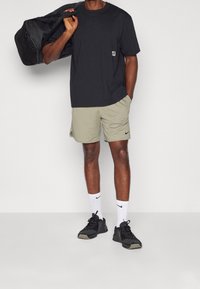 Černé bavlněné tričko s malým logem, béžové sportovní šortky s bočními kapsami, bílé ponožky Nike a černé sportovní boty. Nese černou sportovní tašku.