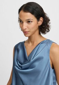 Satin mouwloos topje in zachtblauw met een gedrapeerde colhals en een gladde textuur. Heeft een ontspannen pasvorm en een minimalistisch ontwerp.