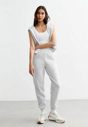 Lichtgrijze joggings met elastische boorden en een elastische tailleband, gecombineerd met een witte tanktop en een grijze mouwloze hoodie. Witte en beige sneakers.
