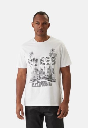 Mężczyzna w białej koszulce z czarnym linearnym rysunkiem palm, panoramy miasta oraz napisem "GUESS Los Angeles CALIFORNIA", w połączeniu z ciemnymi spodniami.