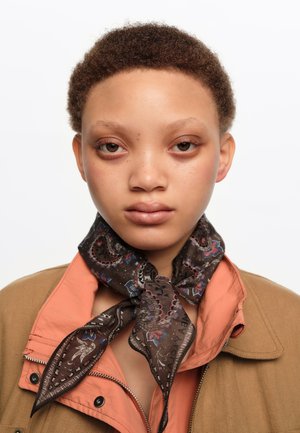 Persona joven con cabello corto y rizado que lleva una chaqueta marrón, cuello color melocotón y una bufanda oscura con estampado atada en el cuello sobre un fondo blanco.