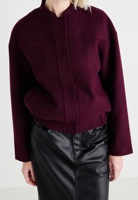 Bordeaux verkorte jacket in gestructureerde stof met een staande kraag en frontnaaddetails, gecombineerd met een zwarte leren kokerrok.