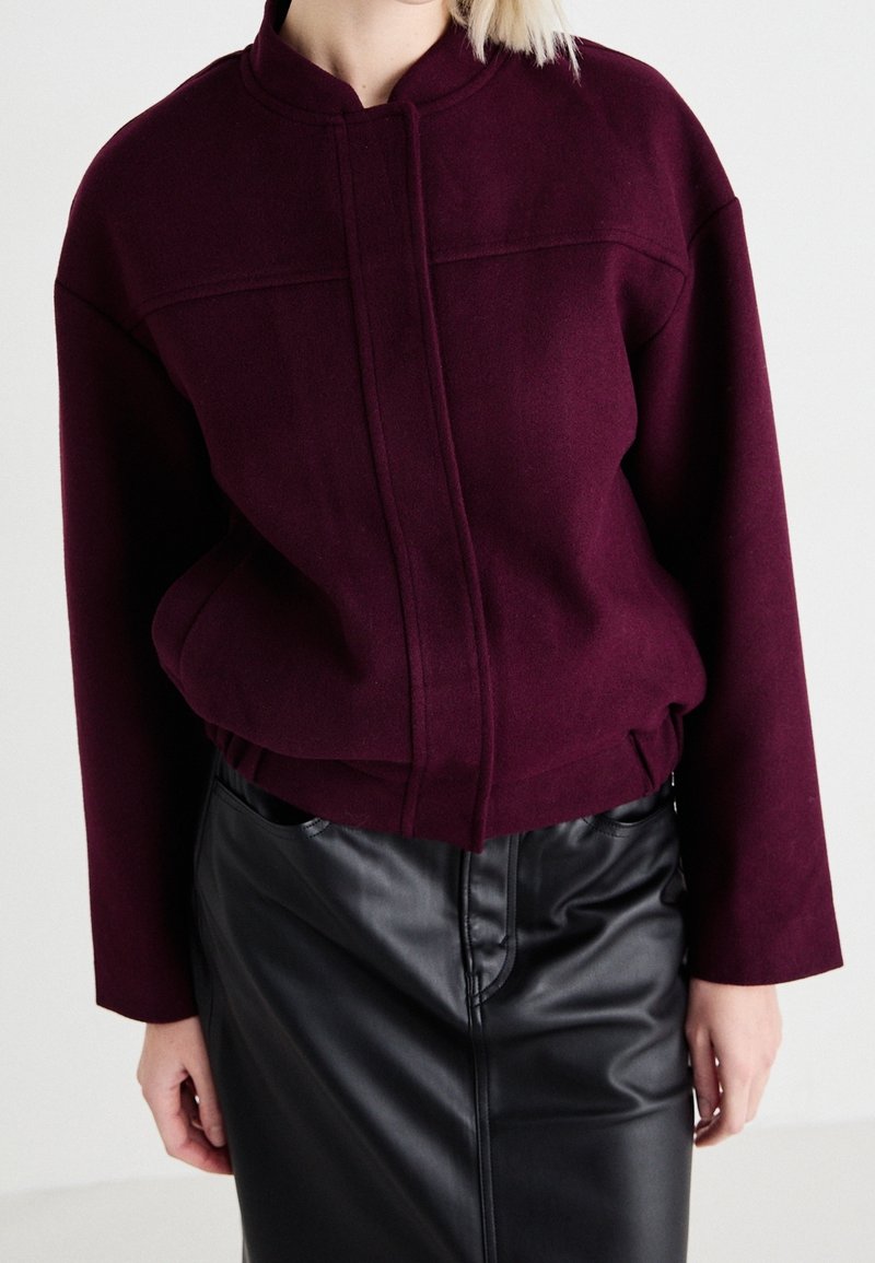 Bordeaux verkorte jacket in gestructureerde stof met een staande kraag en frontnaaddetails, gecombineerd met een zwarte leren kokerrok.