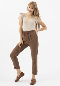 Haut beige à côtes avec décolleté en V et liens aux épaules ; pantalon marron à taille élastique avec cordon de serrage et bas de pantalon resserrés ; mocassins marron avec accents dorés.