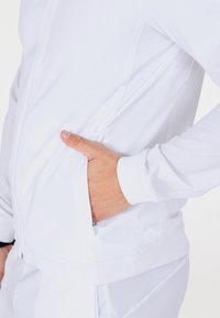 Hånd inde i sidelommen på en hvid zip-up jakke iført matchende hvide bukser mod en neutral baggrund.