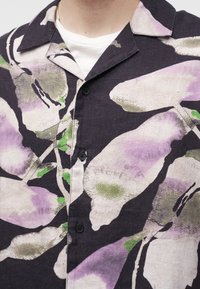 Chemise noire boutonnée avec un large col présentant de grands imprimés floraux abstraits en violet, vert et blanc sur un sous-vêtement blanc.