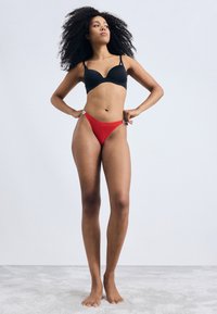 Zwarte gewatteerde beha en rode bikini onderkant op een model, met gladde stof, verstelbare bandjes en een minimalistisch ontwerp.