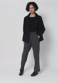 Manteau noir sur mesure porté sur un haut noir, associé à un pantalon en laine gris. Le look est complété par des chaussures noires et une silhouette épurée et moderne.