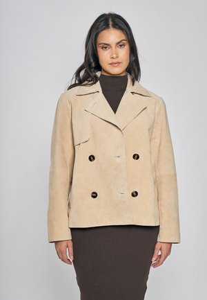 Giacca doppiopetto in camoscio beige con collo a lancia, grandi bottoni scuri e tasche laterali, caratterizzata da una texture liscia e una vestibilità rilassata.