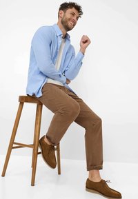 Camicia button-up azzurro chiaro sopra una t-shirt bianca, chinos marroni e scarpe in suede marroni, seduto su uno sgabello di legno contro uno sfondo semplice.