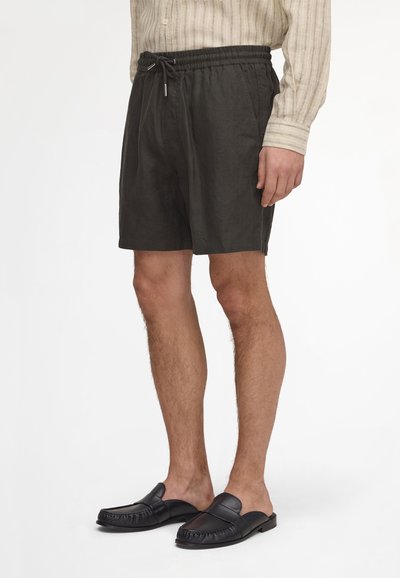Homme portant un short noir à cordon, des mocassins noirs en cuir sans lacets et une chemise beige rayée à manches longues, debout sur un fond blanc.