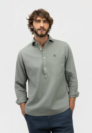 Hombre con cabello rizado y barba, vistiendo una camisa verde de manga larga con botones, de pie con las manos en los bolsillos frente a un fondo liso.