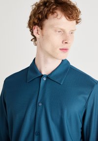 Teal shirt with a button-up design, prominent ribbed texture, and a pointed collar. Features large buttons and smooth fabric finish.