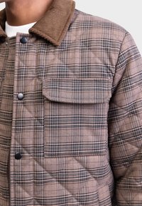 Gros plan d'une veste matelassée à carreaux marron et noir avec un col en velours côtelé marron et des boutons-pression noirs sur la poche avant et la patte de boutonnage.