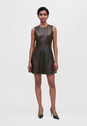 Une femme porte une mini-robe foncée sans manches en similicuir et des talons noirs, sur un fond clair et uni.