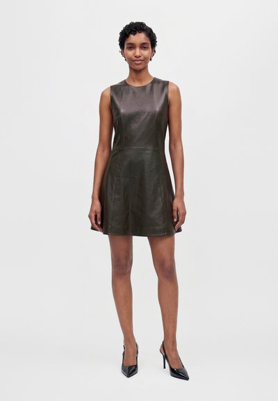 Une femme porte une mini-robe foncée sans manches en similicuir et des talons noirs, sur un fond clair et uni.