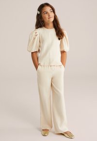Jauna meitene, tērpusies krēmkrāsas puff-sleeve blūzē un atbilstošās platās biksēs, ar zeltītām kurpēm, stāv ar rokām kabatās, skatoties pa labi.