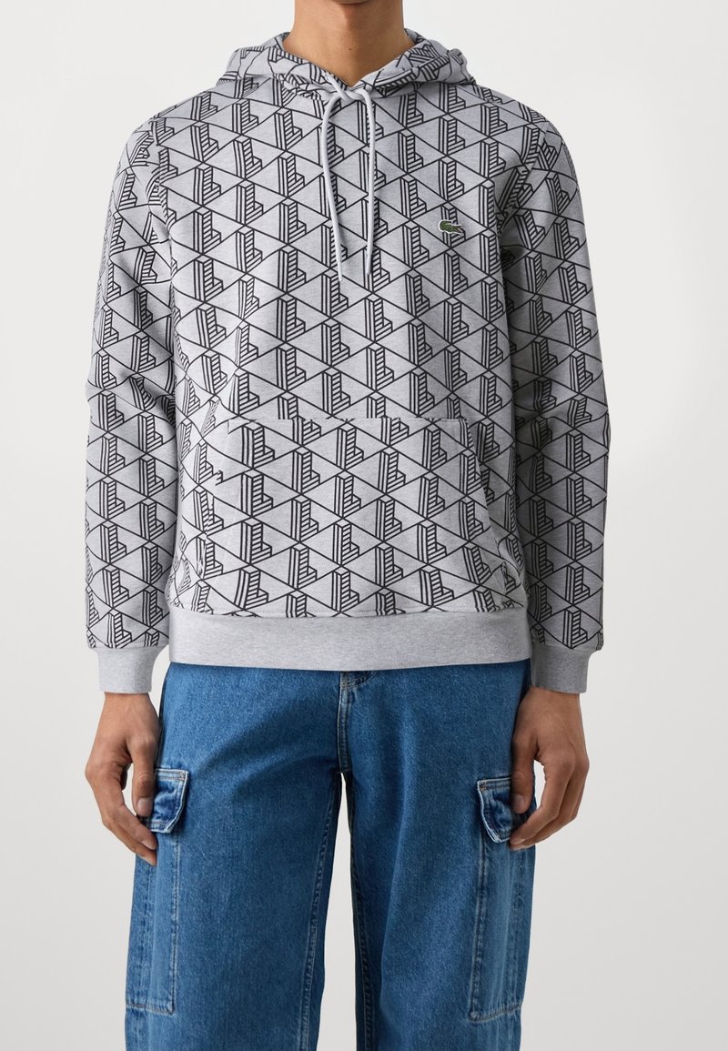 Homme portant un sweat à capuche gris avec un motif géométrique noir et un pantalon cargo en denim bleu sur fond blanc.