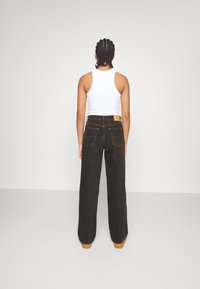 Wijd-uitlopende bruine jeans met een hoge taille, zichtbare steken en twee achterzakken. Gecombineerd met een strakke witte tanktop.