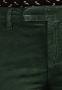 Donkergroene corduroy broek met band en riemlussen, gedeeltelijk ingestoken patroon zwart shirt met witte en rode details.