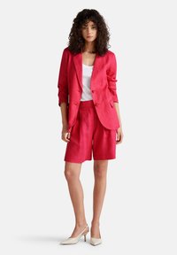 Růžový blazer ze lnu s dvěma knoflíky a odpovídajícími přizpůsobenými šortkami. Modelka má na sobě bílé triko a béžové vysoké podpatky.