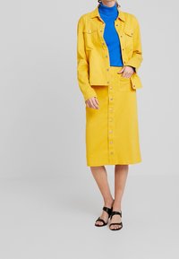 Keltainen farkkutakki ja hameyhdistelmä, jossa on nappikiinnitys, raidalliset taskut ja sininen poolopaita alla. Mustat sandaalit täydentävät asun.