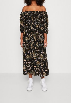 Femme portant une robe mi-longue noire à fleurs avec épaules dénudées, corsage froncé et manches bouffantes, associée à des baskets blanches montantes.