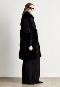 Melnas ādas apmetnis ar dažādām tekstūrām, tajā skaitā pūkains un gluds audums, augsts apkakles dizains un platais piedurknes, kombinēts ar vaļīgiem melniem biksēm.