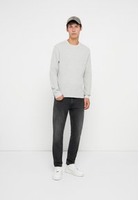 Svetlošedý rebrovaný sveter, čierne slim-fit džínsy a biele tenisky. Model nosí šedú čiapku a stojí proti bielej stene.