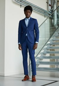 Homme en costume bleu et chemise bleu clair se tenant à l'intérieur près d'un escalier en métal avec balustrade en verre, portant des chaussures habillées marron.