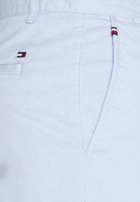 Weiße Baumwollhosen mit einer Fronttasche, die ein gesticktes Logo in Rot, Weiß und Blau aufweist. Glatte Textur, taillierte Passform und sichtbare Nähte.