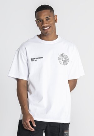 Usmievavý muž v bielom oversized tričku s nápisom "Gianni Kavanagh Since 2017" a kruhovým logom, čiernych nohaviciach, stojaci pred jednoduchým pozadím.