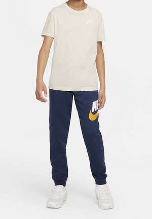 Teenager iført beige Nike T-shirt med korte ærmer, marineblå joggingbukser med stor gul Nike-swoosh og hvide sneakers, stående mod en hvid baggrund.
