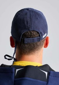 Cappellino in cotone blu navy con visiera curva, cinturino regolabile sul retro e dettagli del logo. Camicia gialla e colletto visibili sotto.