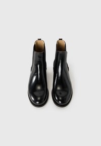 Une paire de bottines Chelsea en cuir verni noir avec doublure intérieure beige, panneaux latéraux élastiques et tirettes sur fond blanc.