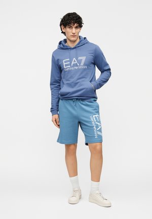 Modrá bavlnená mikina s kapucňou s logom EA7, doplnená o modré šortky s bielym potlačeným logom. Model má na nohách béžové tenisky a biele ponožky.