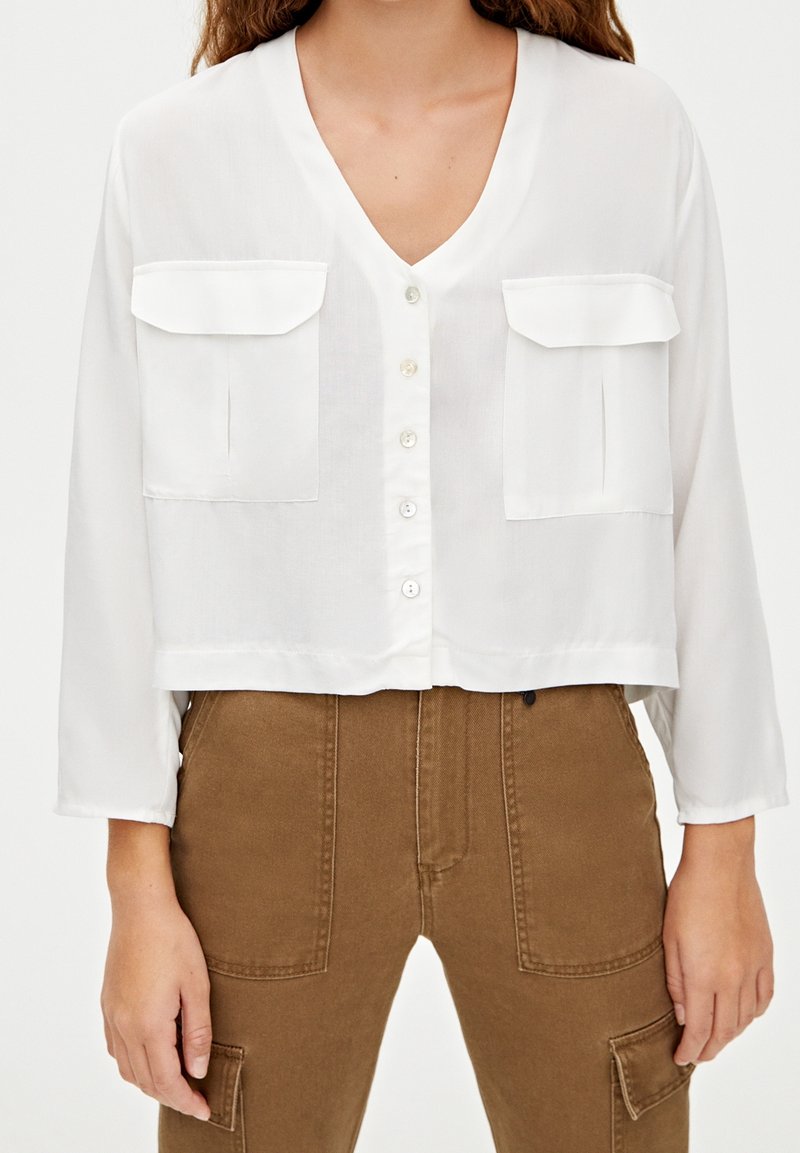Brassière blanche coupée avec un col en V, des manches longues et deux poches poitrine. Fabriquée en tissu léger avec un devant à boutons.