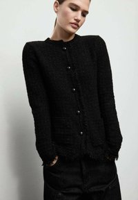 Czarny sweter o fakturze z okrągłym dekoltem, długimi rękawami i zapięciem na guziki z przodu, z postrzępionymi krawędziami wzdłuż dołu.
