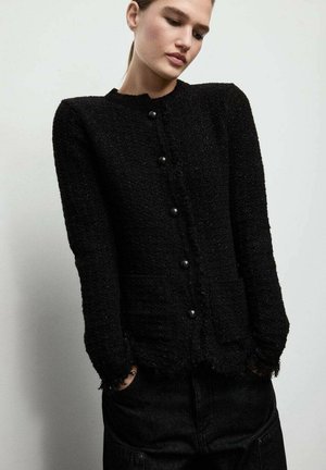 Cardigan noir texturé avec un col rond, des manches longues et des fermetures à boutons à l'avant, présentant des bords effilochés le long de l'ourlet.