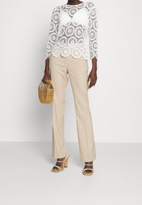 Haut en dentelle blanche avec des motifs circulaires, pantalon beige droit, sandales à talons croisées couleur fauve et sac en osier.