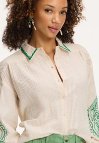 Camicia bianca con colletto, bottoni e rifiniture decorative verdi, in tessuto leggermente texturizzato, con ricami sulle maniche. Abbinata a pantaloni verdi in tessuto texturizzato.