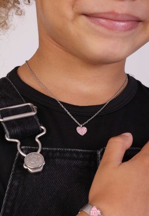 Zilveren ketting met een kleine roze harthanger, heeft gestructureerde details en is omgeven door een metalen rand. Gedeeltelijk zichtbaar boven een zwarte top.