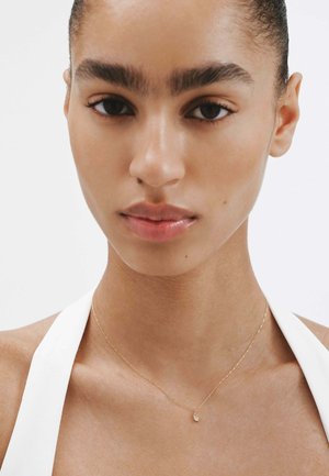 Collar de oro en un cuello desnudo, con un pequeño colgante. Textura de piel suave, tono marrón claro y maquillaje mínimo, contra un fondo blanco.
