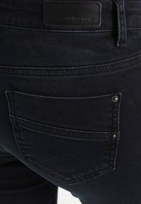 Tmavě modré džíny z denimu s zadní kapsou, která je ozdobena stříbrnými nýty. Tkanina má hladkou strukturu a je viditelný černý kožený štítek značky.