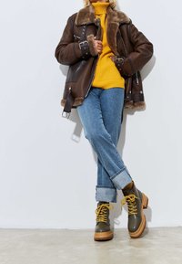 Chaqueta de bomber de piel de oveja marrón, suéter de cuello alto amarillo, jeans azul claro con dobladillo y botas verdes con cordones amarillos y suelas marrones.