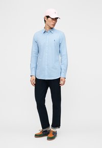 Fiatal férfi világoskék kockás ingben, fekete nadrágban, színes hajócipőben és rózsaszín baseball sapkában, fehér háttér előtt állva.