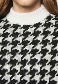 Suéter de punto con un patrón de padrón de houndstooth en blanco y negro, y un cuello blanco acanalado. El material parece suave y texturizado.