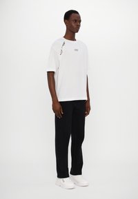 T-shirt blanc à manches courtes avec un détail incurvé noir et blanc sur l'épaule, associé à un pantalon noir et des baskets blanches.