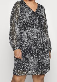 Vestido negro con flores, mangas largas transparentes, escote en forma de v, cintura ceñida y una falda fluida con un patrón de flores blancas.